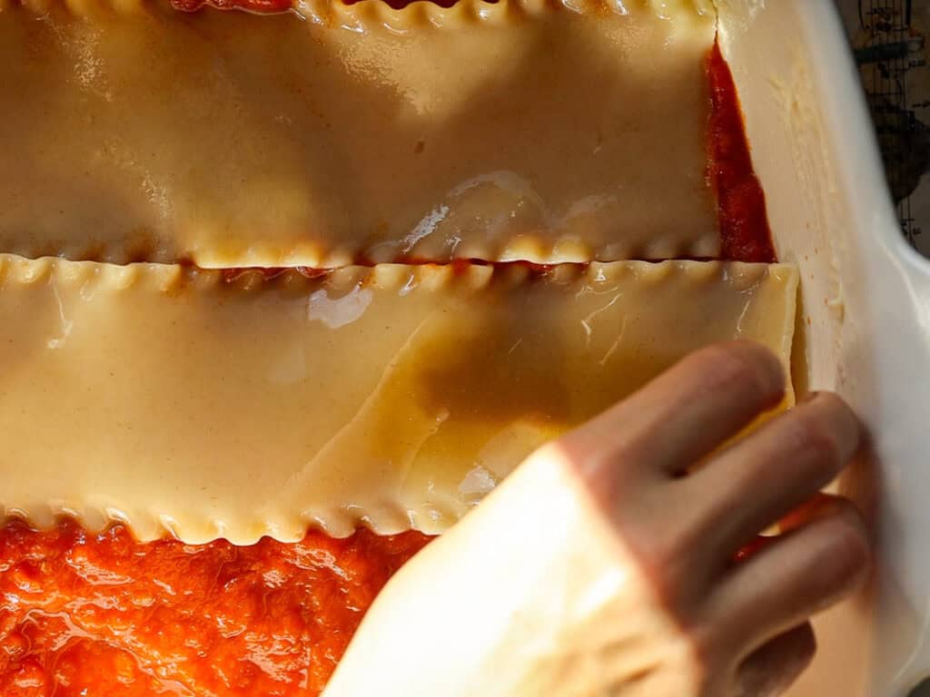 A person assembling a lasagna by layering uncooked lasagna noodles over tomato sauce in a white baking dish. The image is well-lit, highlighting the textures and colors of the pasta and sauce.